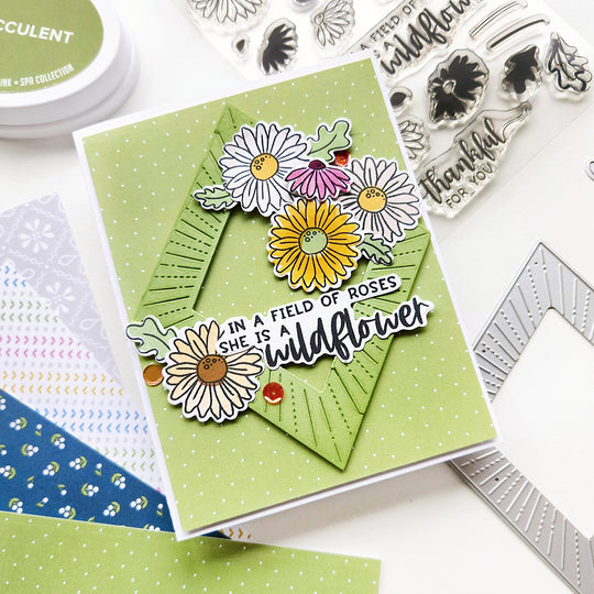 A card with the sentiment "In a field of roses, she is a wildflower," featuring daisy accents on a sunburst diamond shape, using green polka dot patterned paper.