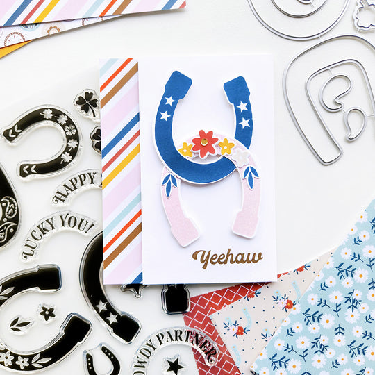 A card featuring double horseshoes adorned with stars and flowers, with a "Yeehaw" sentiment, against stripe patterned paper for a western design.