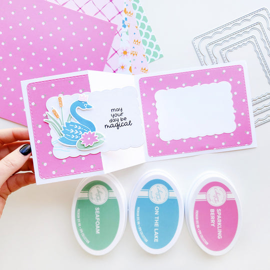 "May your day be magical" card featuring a swan on a rippled pond with cattails and a lily pad, set on floral-patterned paper