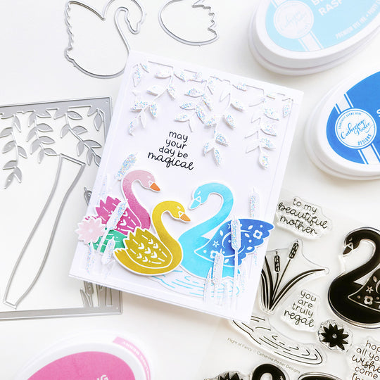 "May your day be magical" card with three swans gliding on a rippled pond, surrounded by cattails, a lily pad, and cascading vines.